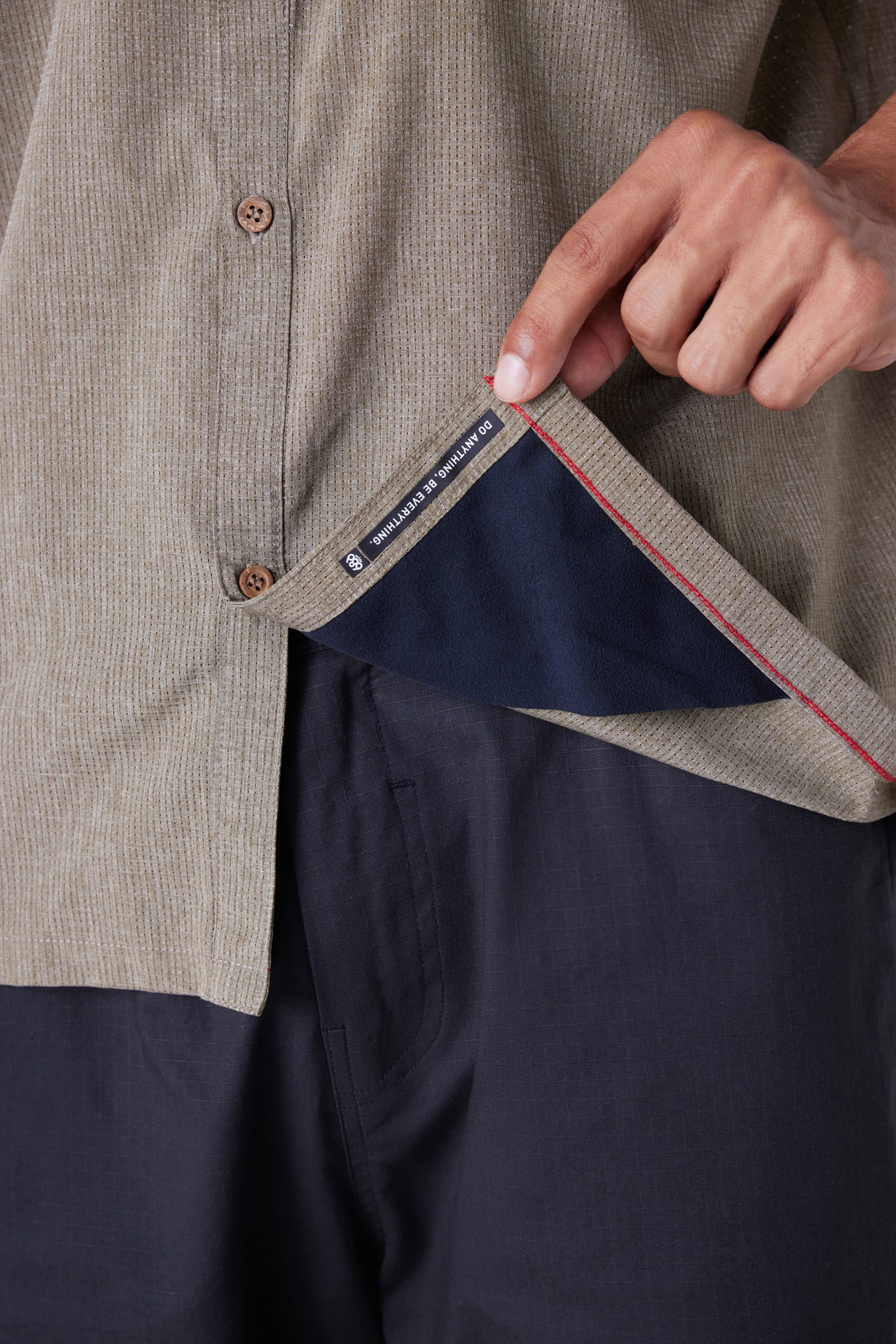 A person lifts the hem of a 686 Mens Seeker Perforated Button Up shirt in beige, revealing a black 686 label with white text inside. They are wearing dark pants, with only their torso and hand visible.