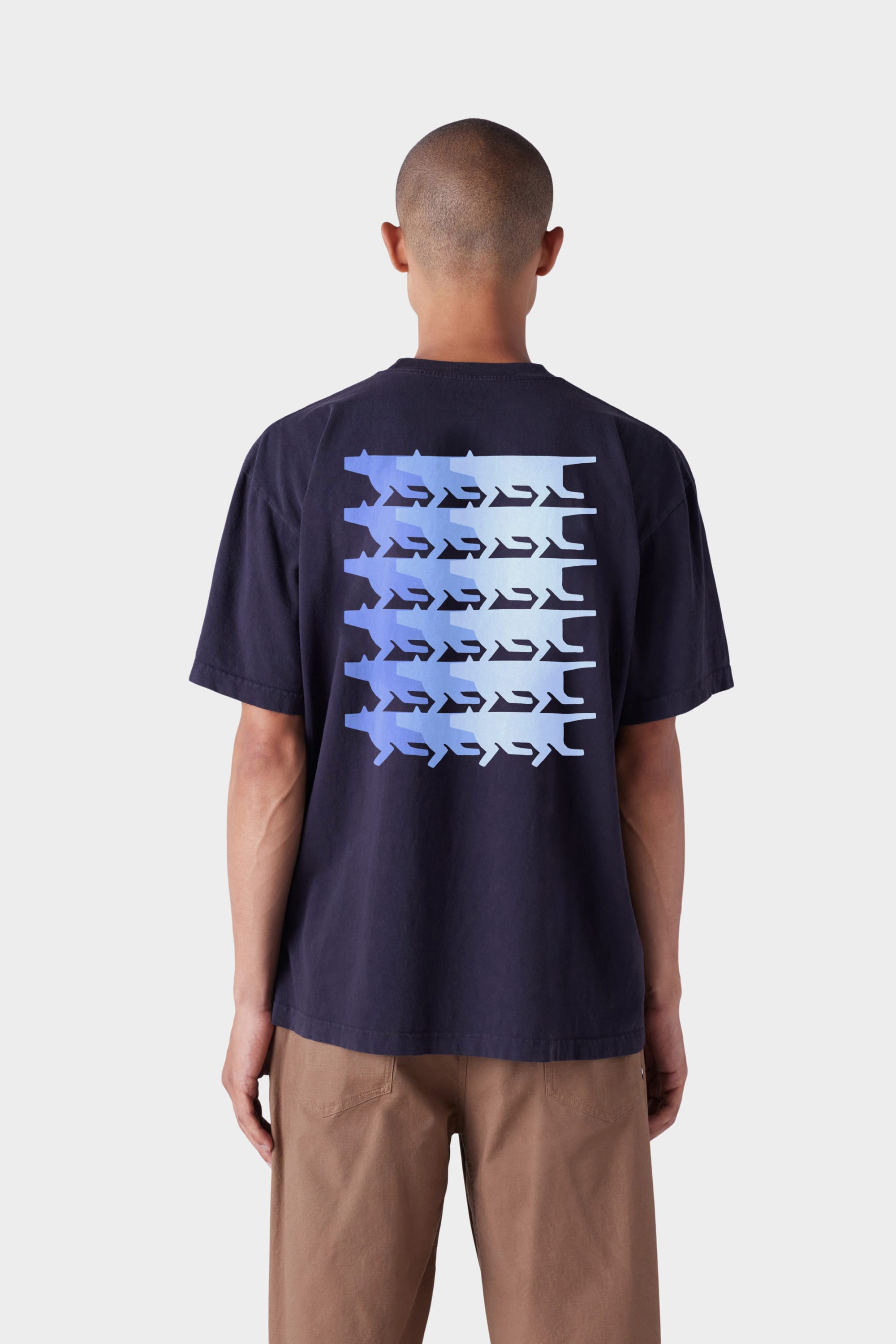 A person wearing the 686 Mens Wanderers Short Sleeve Tee by 686, featuring blue and white shark silhouettes on the back, paired with light brown pants, stands facing away from the camera against a plain background.