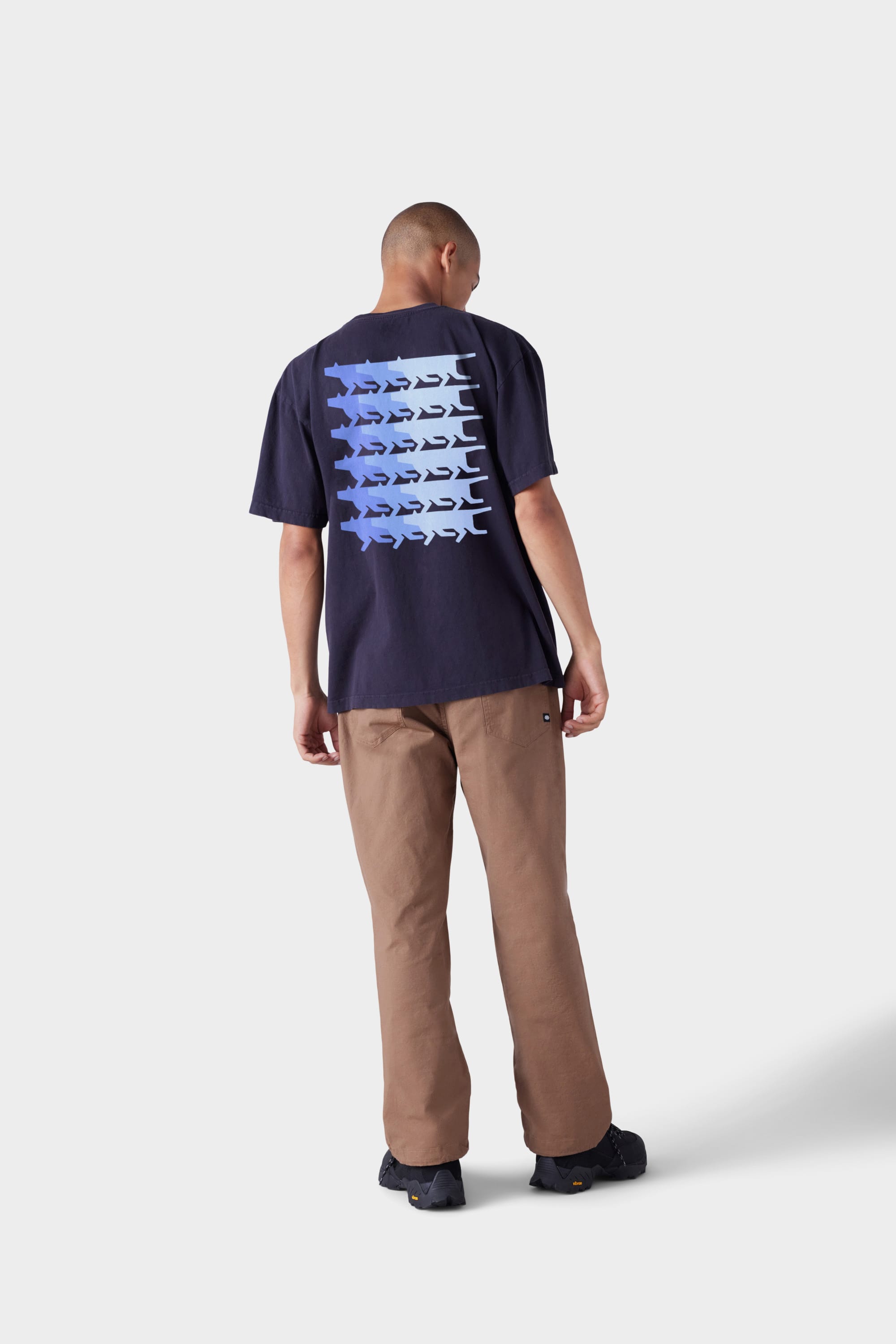 A person stands facing away, wearing the 686 Mens Wanderers Short Sleeve Tee by 686 in black with a blue shark pattern on the back, paired with brown pants and black shoes against a plain light gray background.