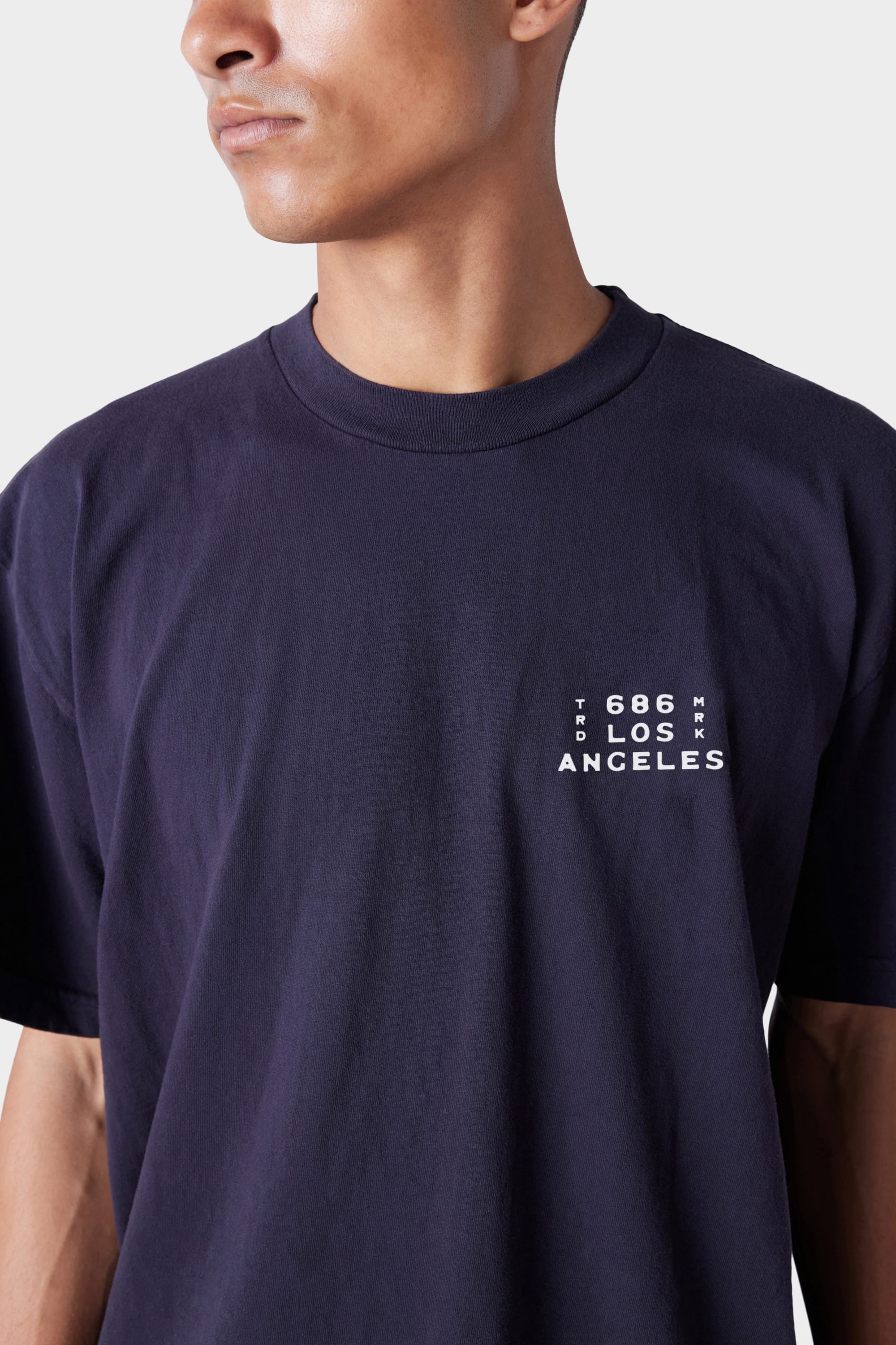 A person wears the 686 Mens Wanderers Short Sleeve Tee by 686, a dark navy shirt featuring small white 606 LOS ANGELES text in a square on the upper left. The background is plain and the persons face is partially out of frame.