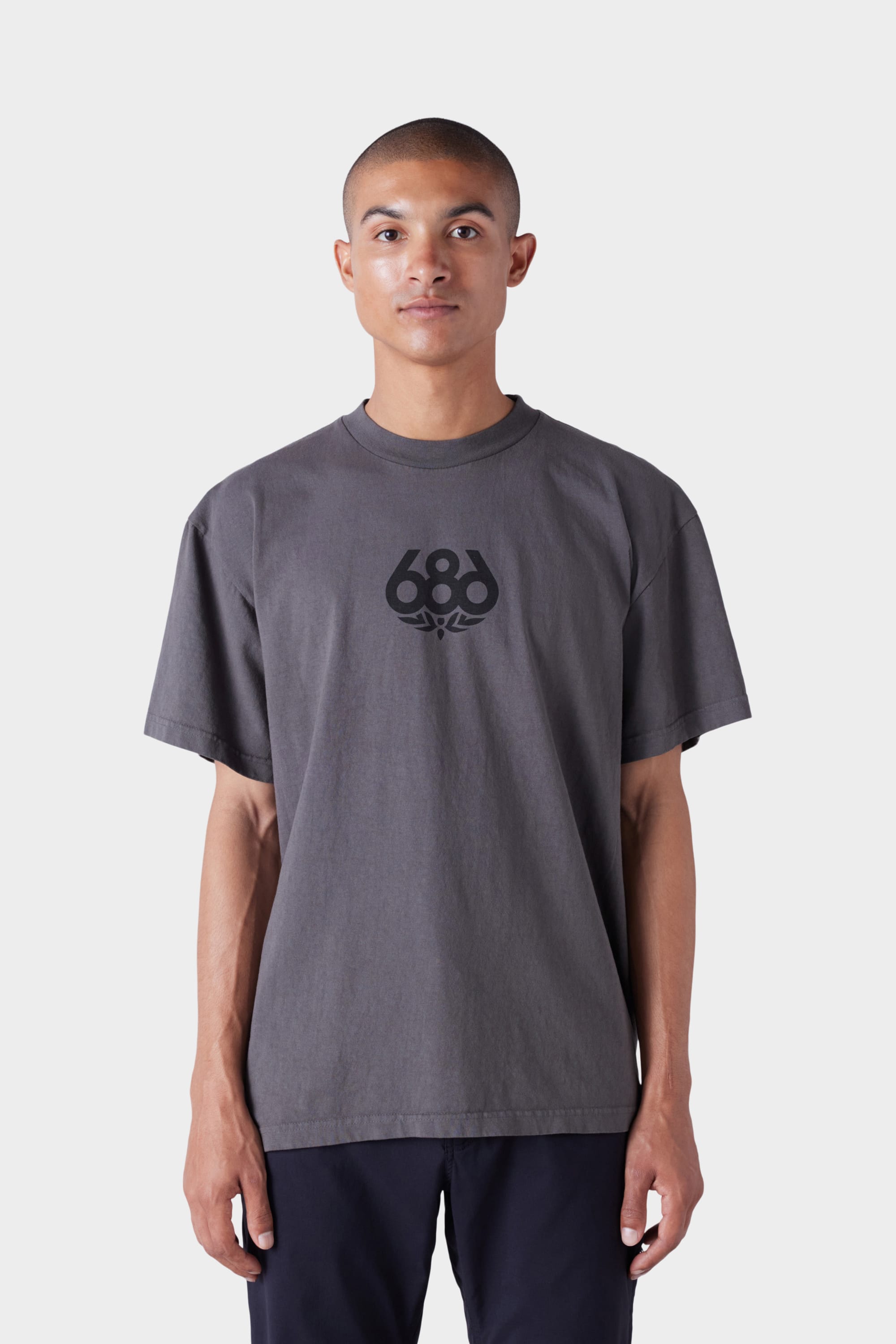 A person with a shaved head wears the 686 Mens Wreath Short Sleeve Tee, a loose-fitting dark gray t-shirt with a black 686 logo on the chest, standing against a plain light background.