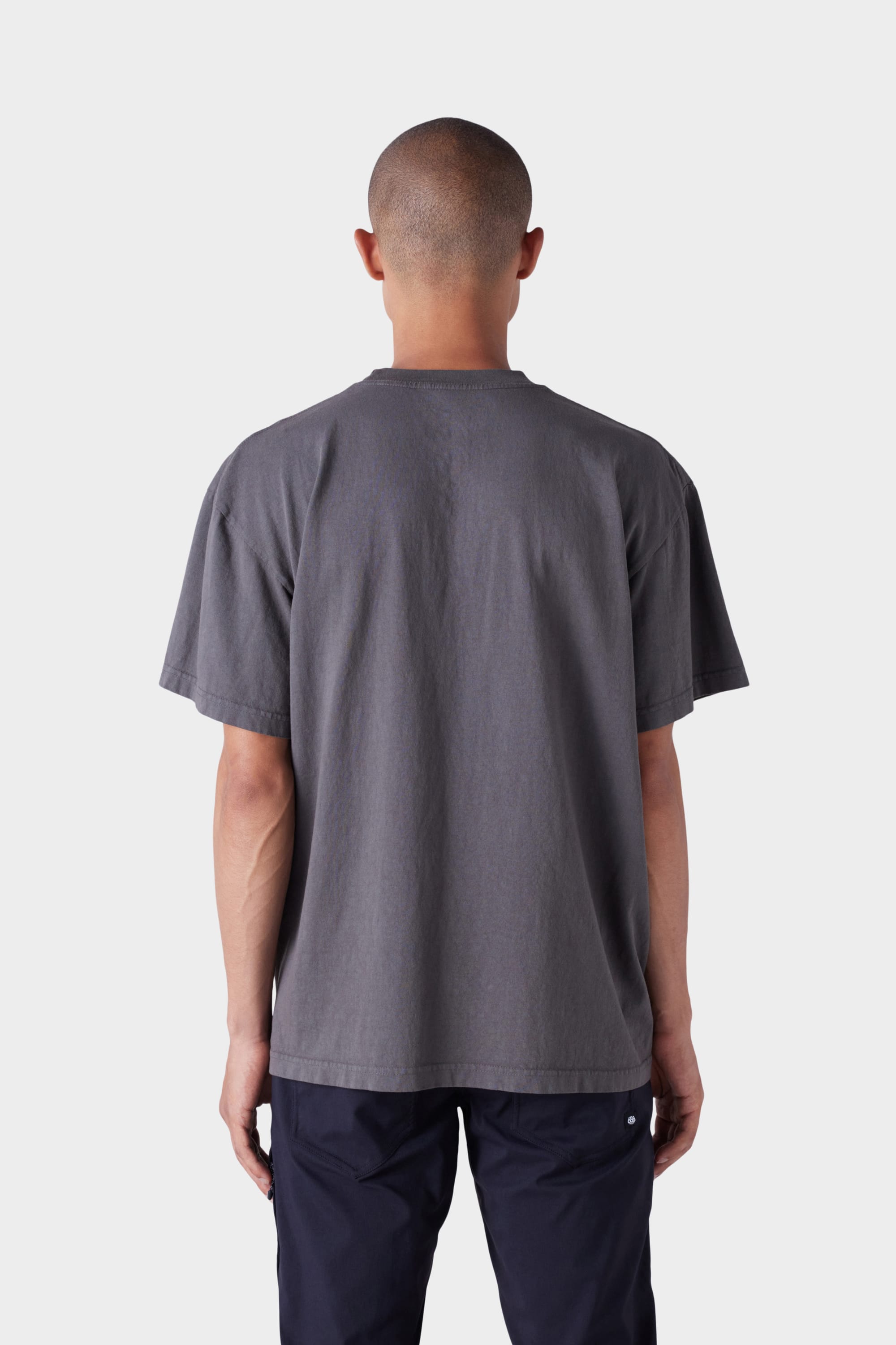 A person with a shaved head stands with their back to the camera, wearing the 686 Mens Wreath Short Sleeve Tee by 686 in dark gray and dark pants against a plain light background.