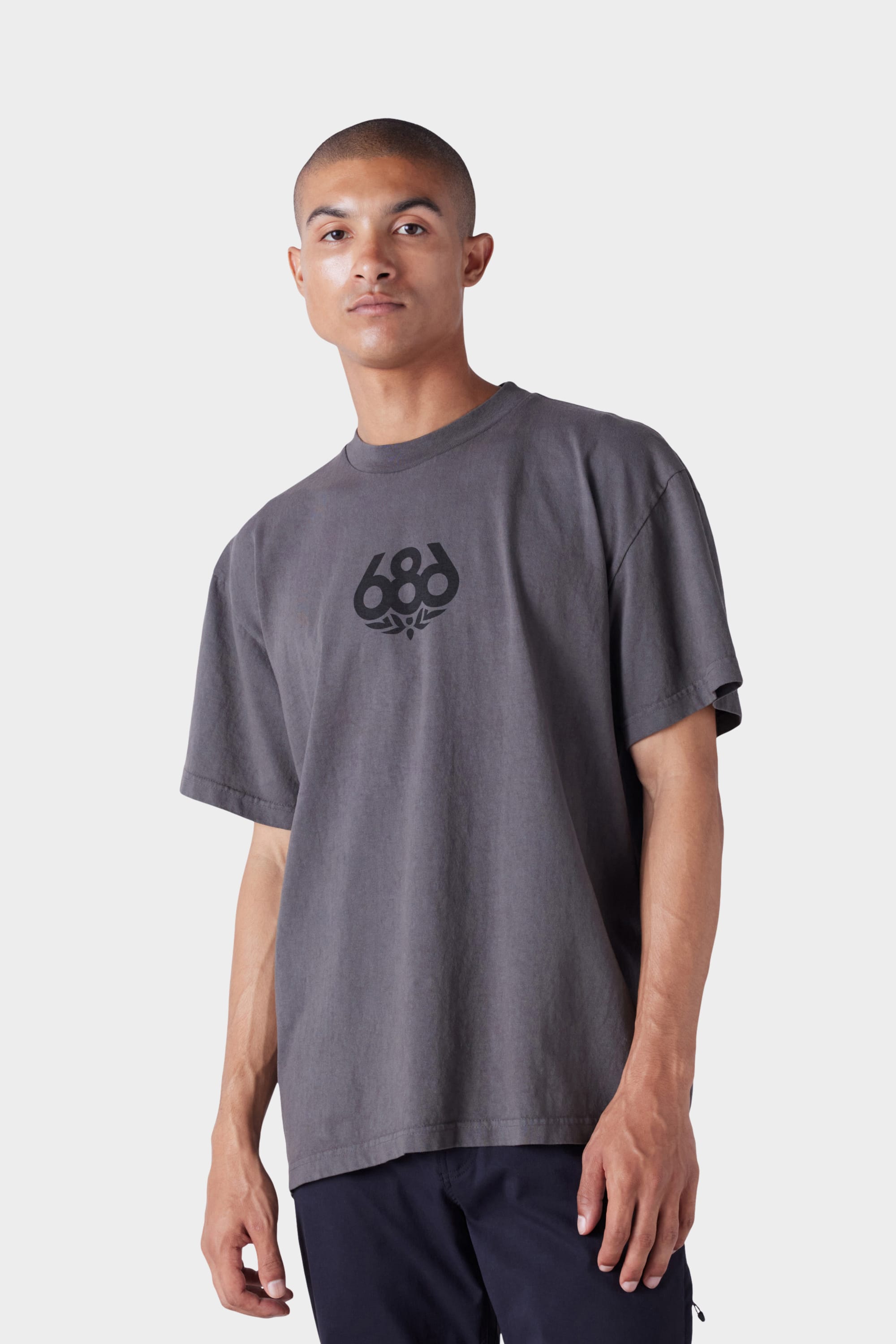 A man with short hair and a neutral expression wears the 686 Mens Wreath Short Sleeve Tee—a loose-fitting, faded gray t-shirt with a black 686 logo on the chest—against a plain light gray background.