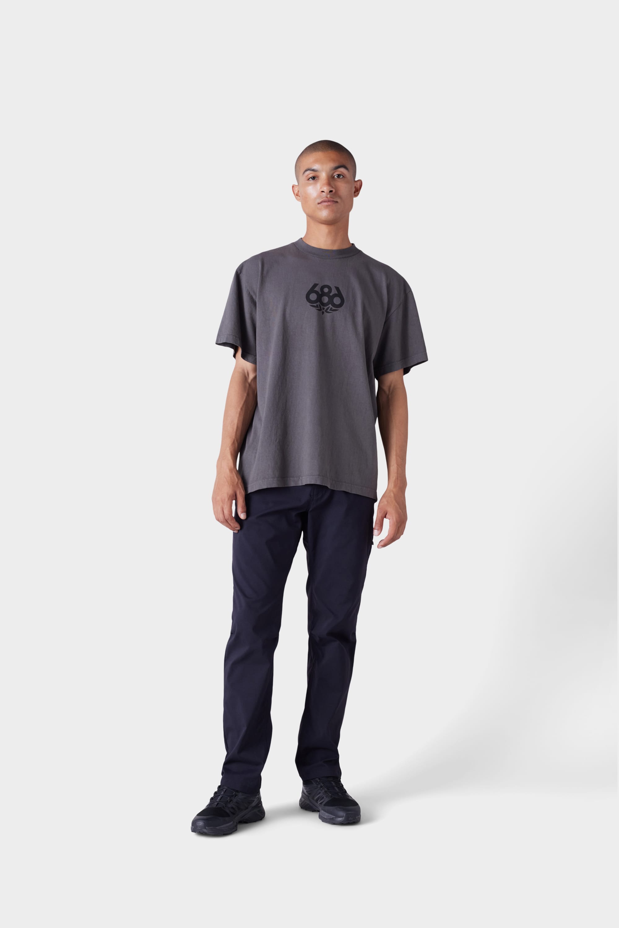 A person with short hair and a neutral expression wears the 686 Mens Wreath Short Sleeve Tee by 686, featuring a loose fit and circular logo in the center, paired with navy pants and black sneakers against a plain white background.