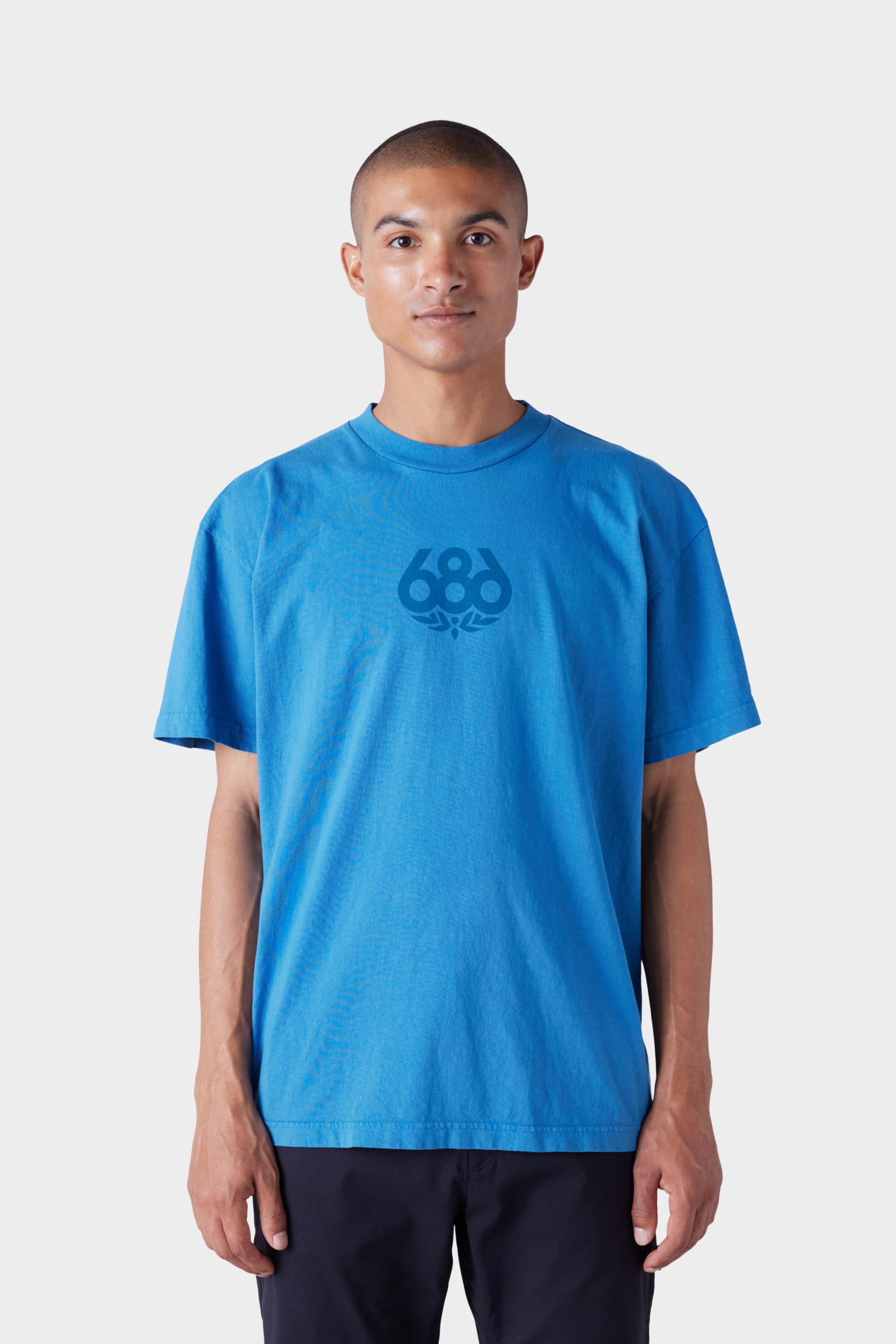 A person with short hair wears the 686 Mens Wreath Short Sleeve Tee—a loose-fitting blue T-shirt by 686 featuring a chest design—while standing facing forward against a plain white background.