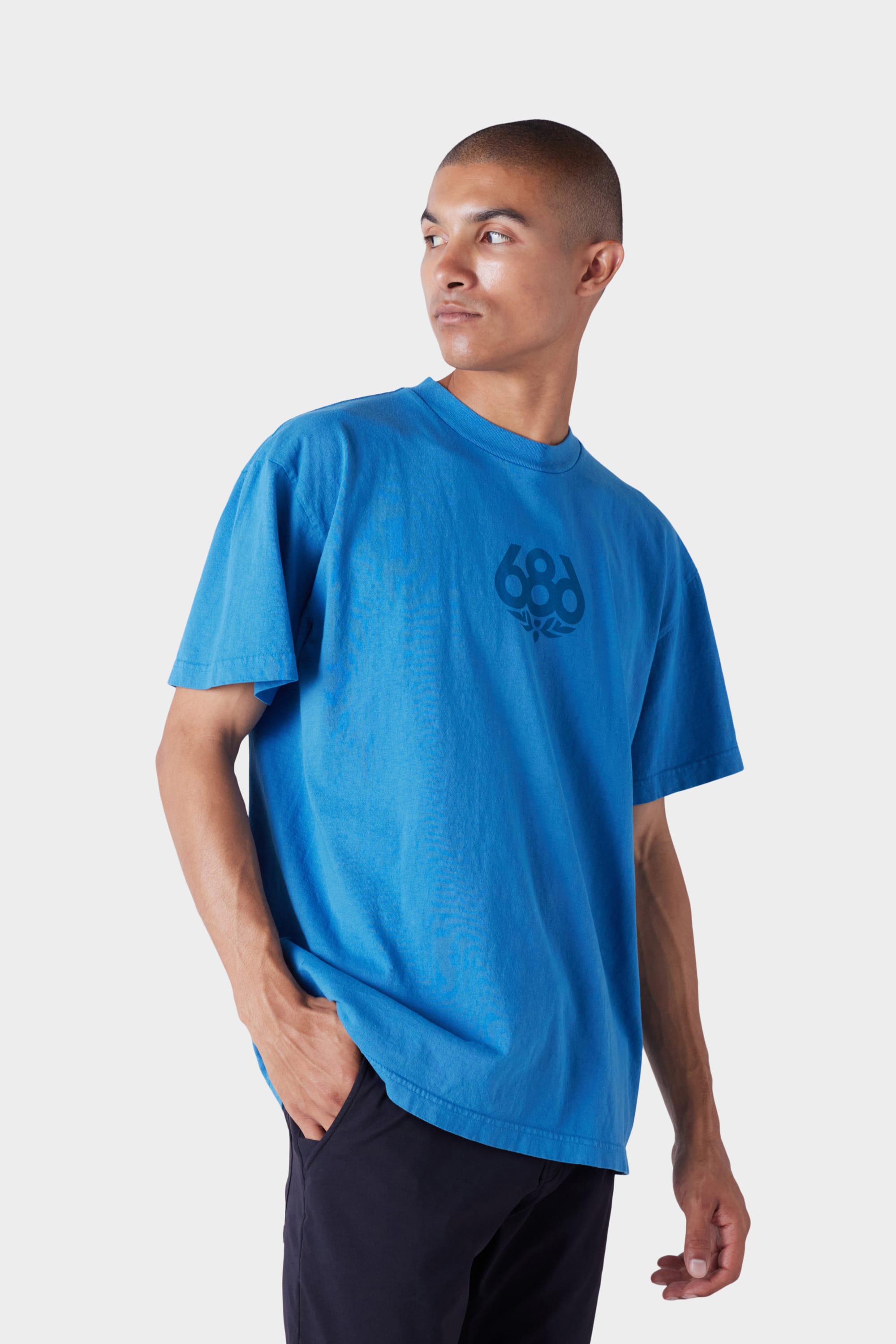 A person in the 686 Mens Wreath Short Sleeve Tee—a loose-fitting blue t-shirt with a 686 logo on the chest—stands against a plain light background, looking left with one hand in their pocket.