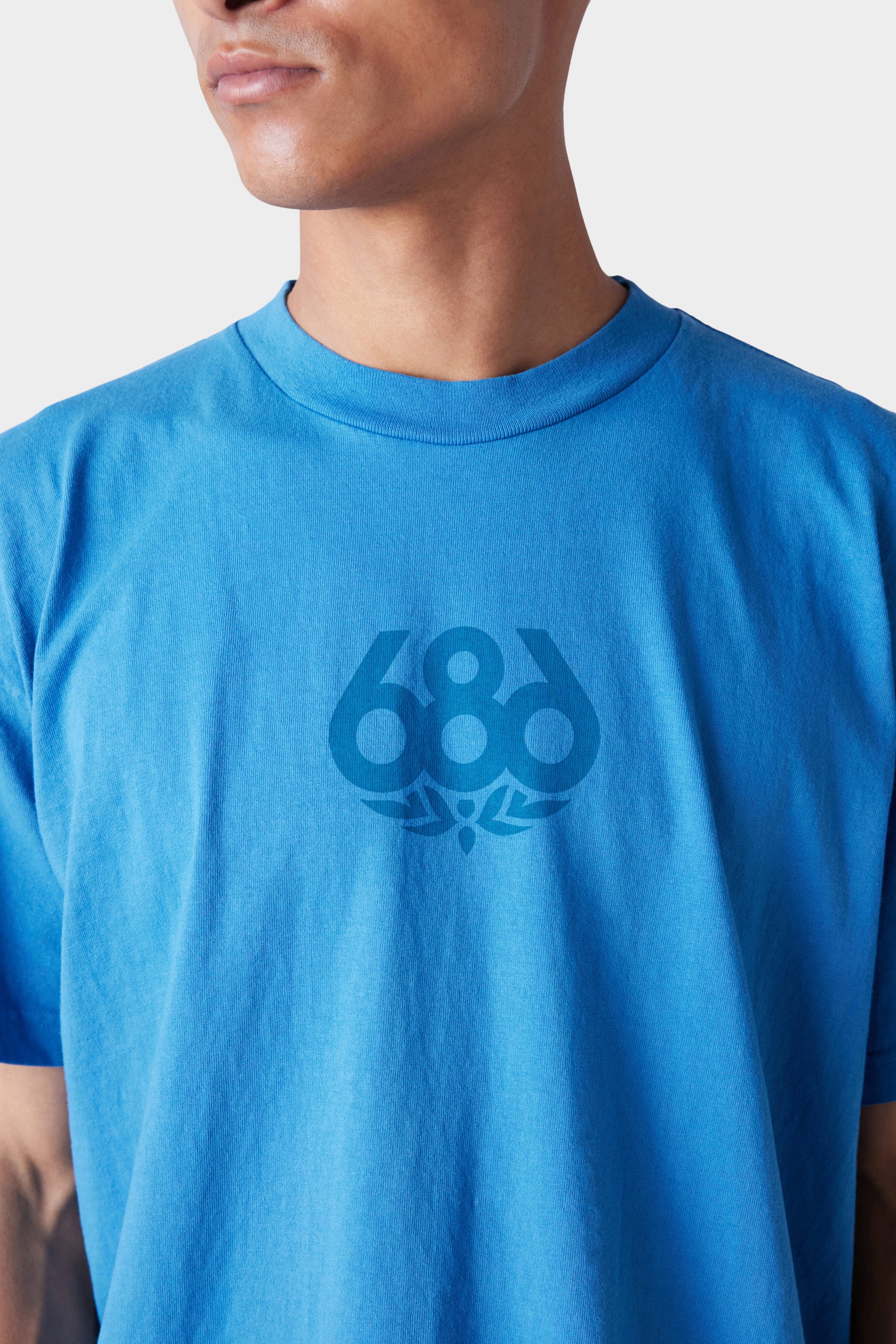 A person is wearing the 686 Mens Wreath Short Sleeve Tee—a bright blue t-shirt featuring a dark blue 686 logo with a small decorative chest design. The background is plain and light-colored.