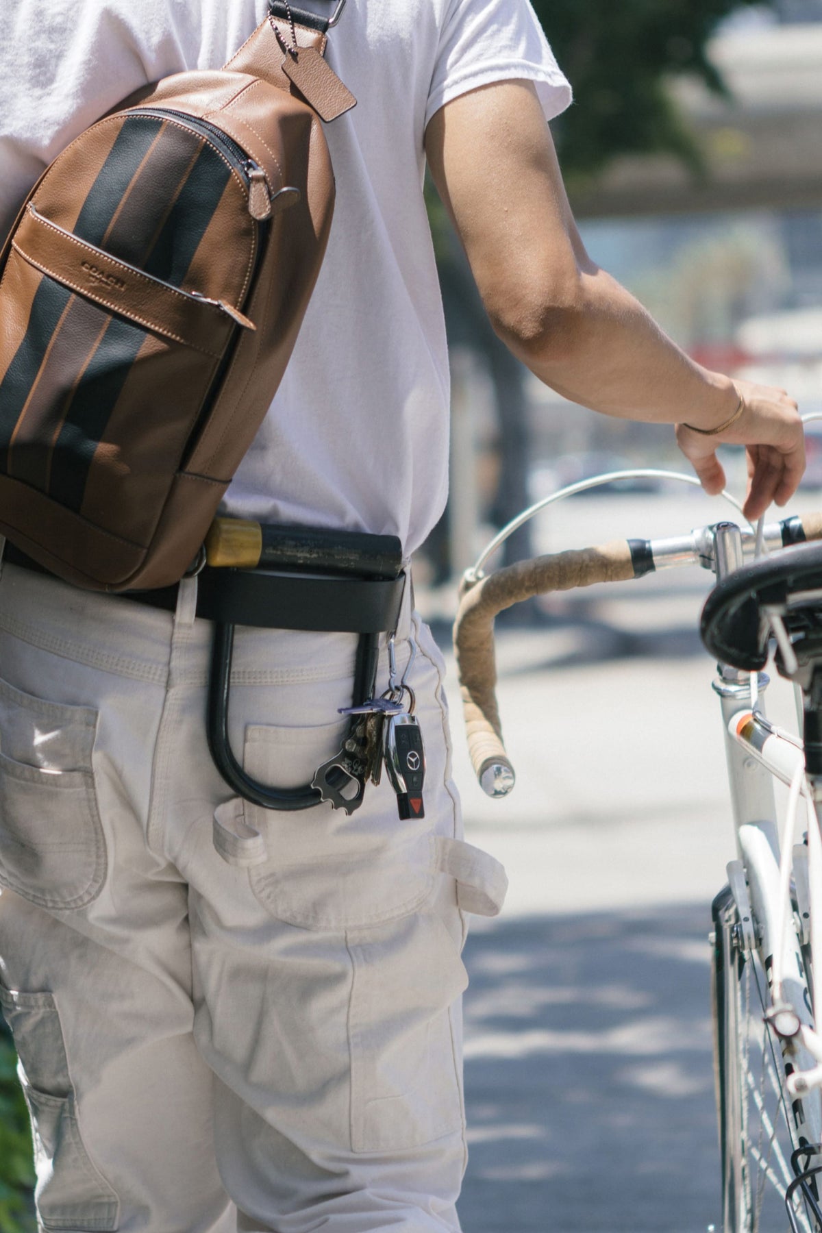A person in white pants and shirt walks a bicycle outdoors on a sunny day, carrying a brown backpack. Keys and the Westwell 686 Westwell Voyager Belt bike lock hang from their belt.