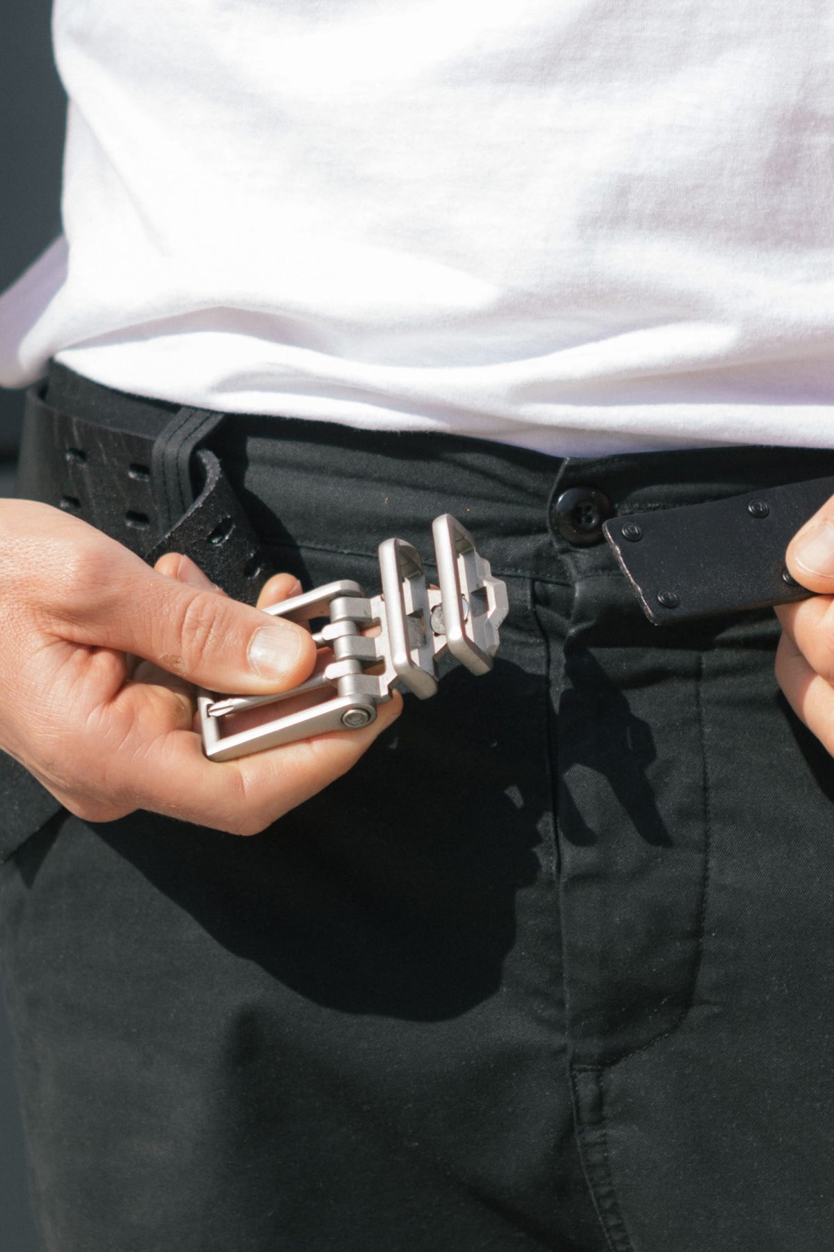 A person wearing black pants and a white shirt is fastening the metallic buckle of the 686 Westwell Voyager Belt by Westwell, preparing to insert it into the belt holes.