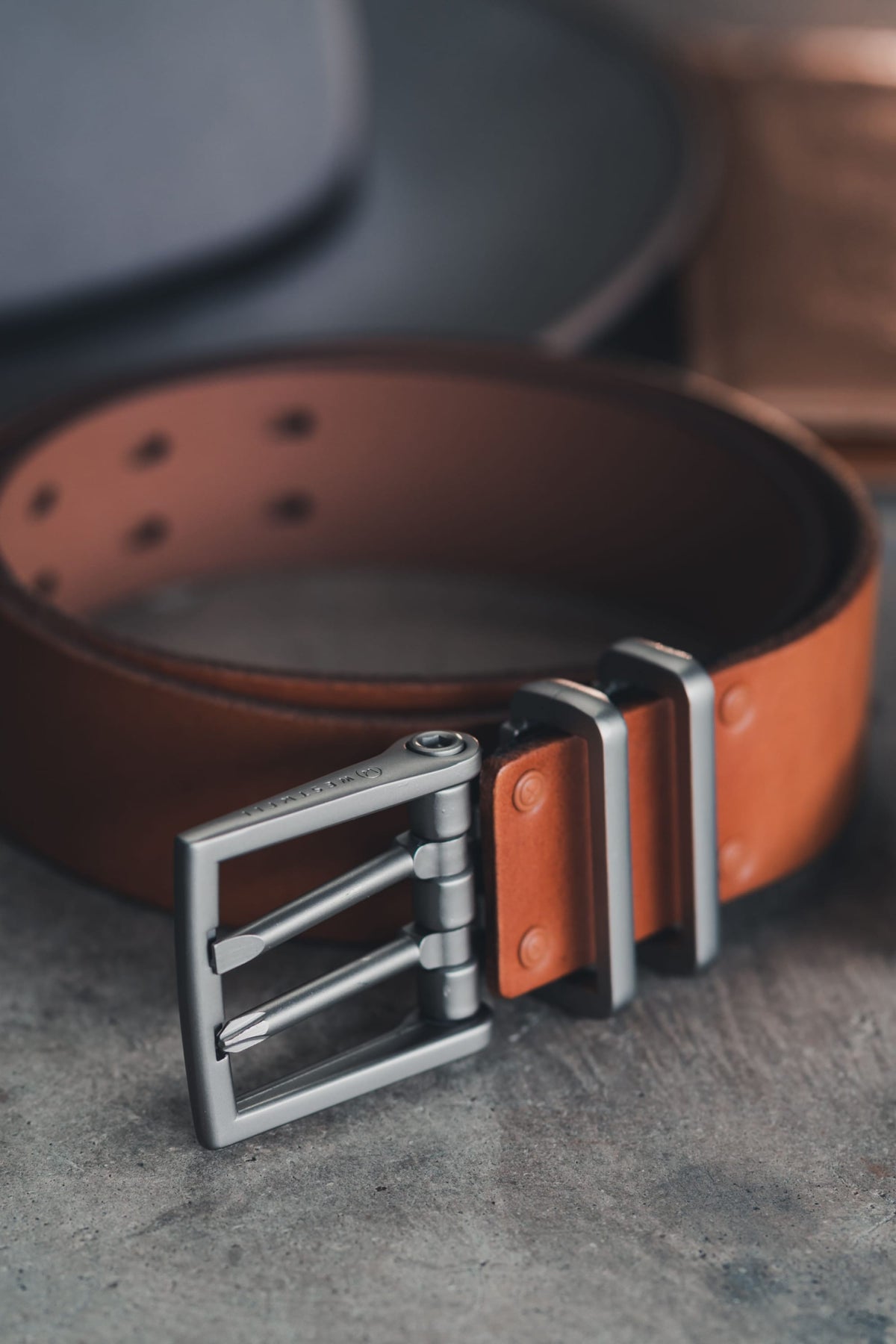 The 686 Westwell Voyager Belt by 686, featuring brown leather and a silver double-prong buckle with metal rivets, is shown coiled on a grey textured surface.