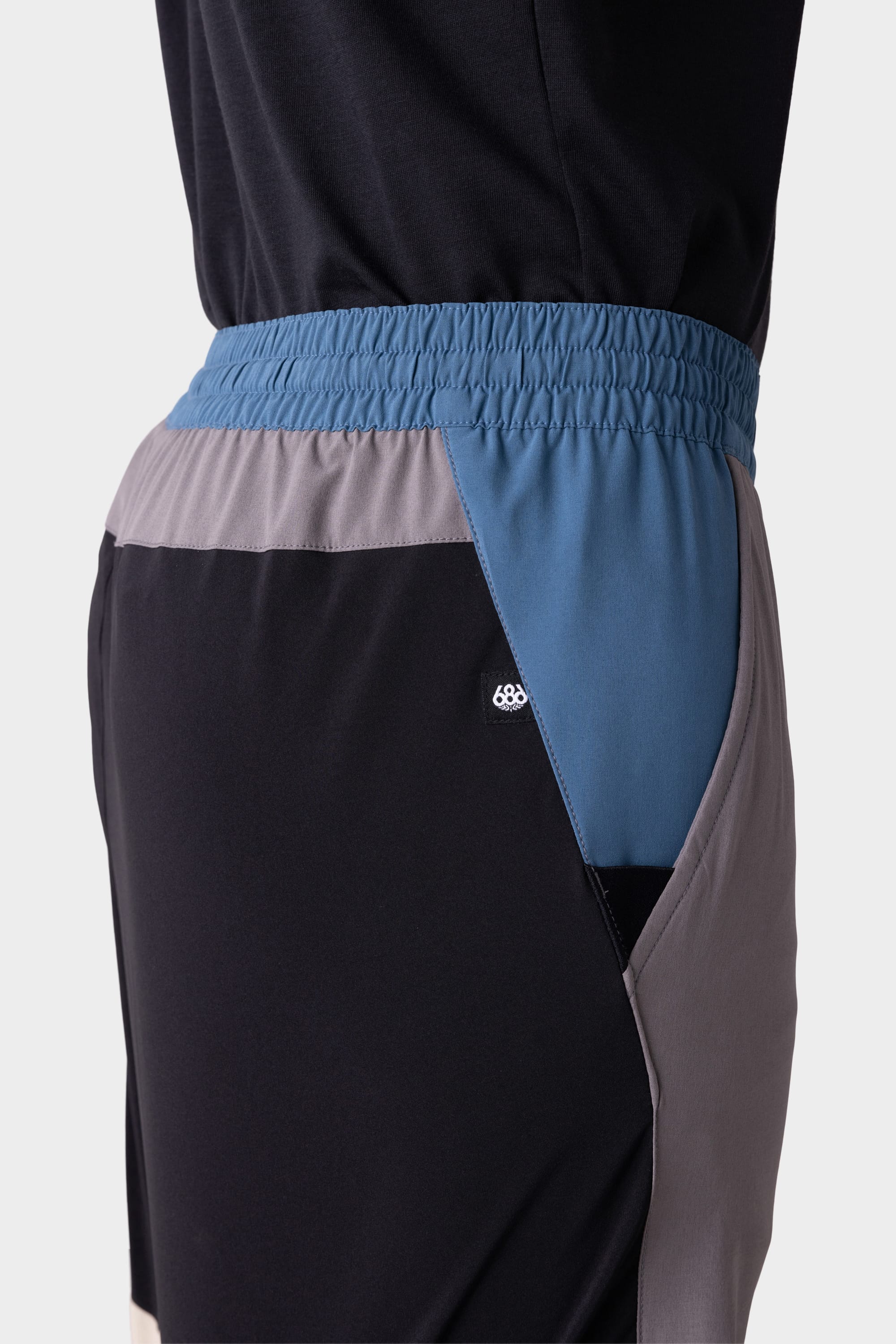 A close-up side view of a person wearing the 686 Mens ATP Stretch Performance Short by 686, featuring blue, grey, and black color-blocked panels, an elastic waistband, and a pocket.