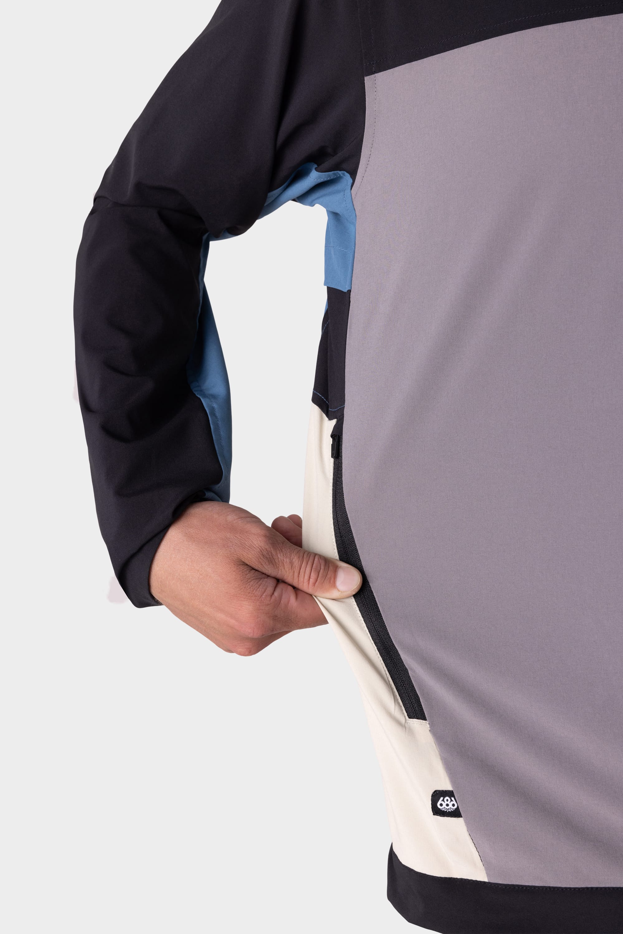A hand unzips a side pocket on the 686 Mens ATP Stretch Wind Jacket, featuring black, grey, blue, and cream color-block sections. Only part of the jacket and hand are visible against a plain light background.