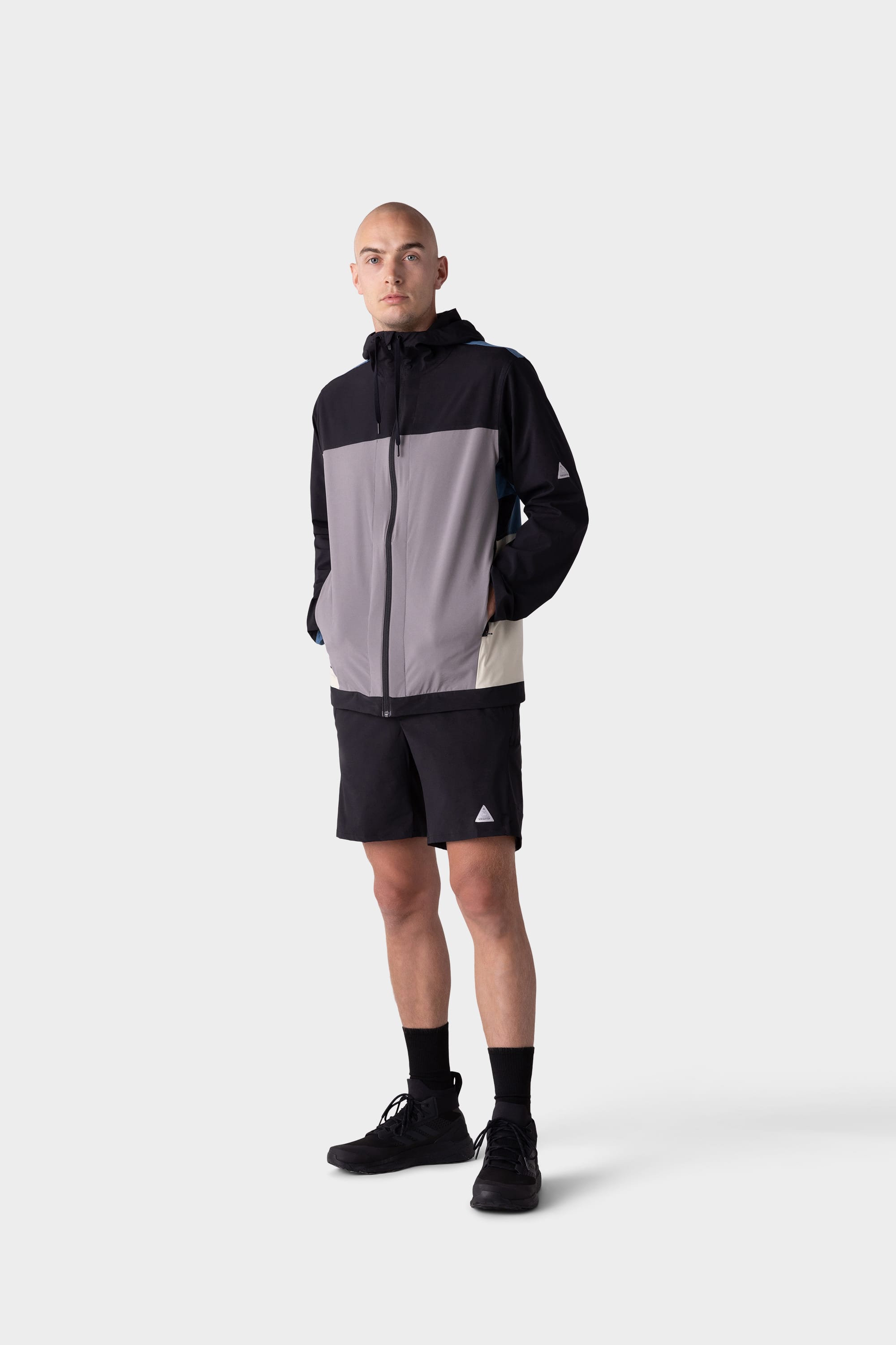 A person stands against a plain light background, wearing the 686 Mens ATP Stretch Wind Jacket by 686 in black and gray color-blocks, paired with black shorts, socks, and sneakers. Their hands are in the jacket pockets and they have a neutral expression.