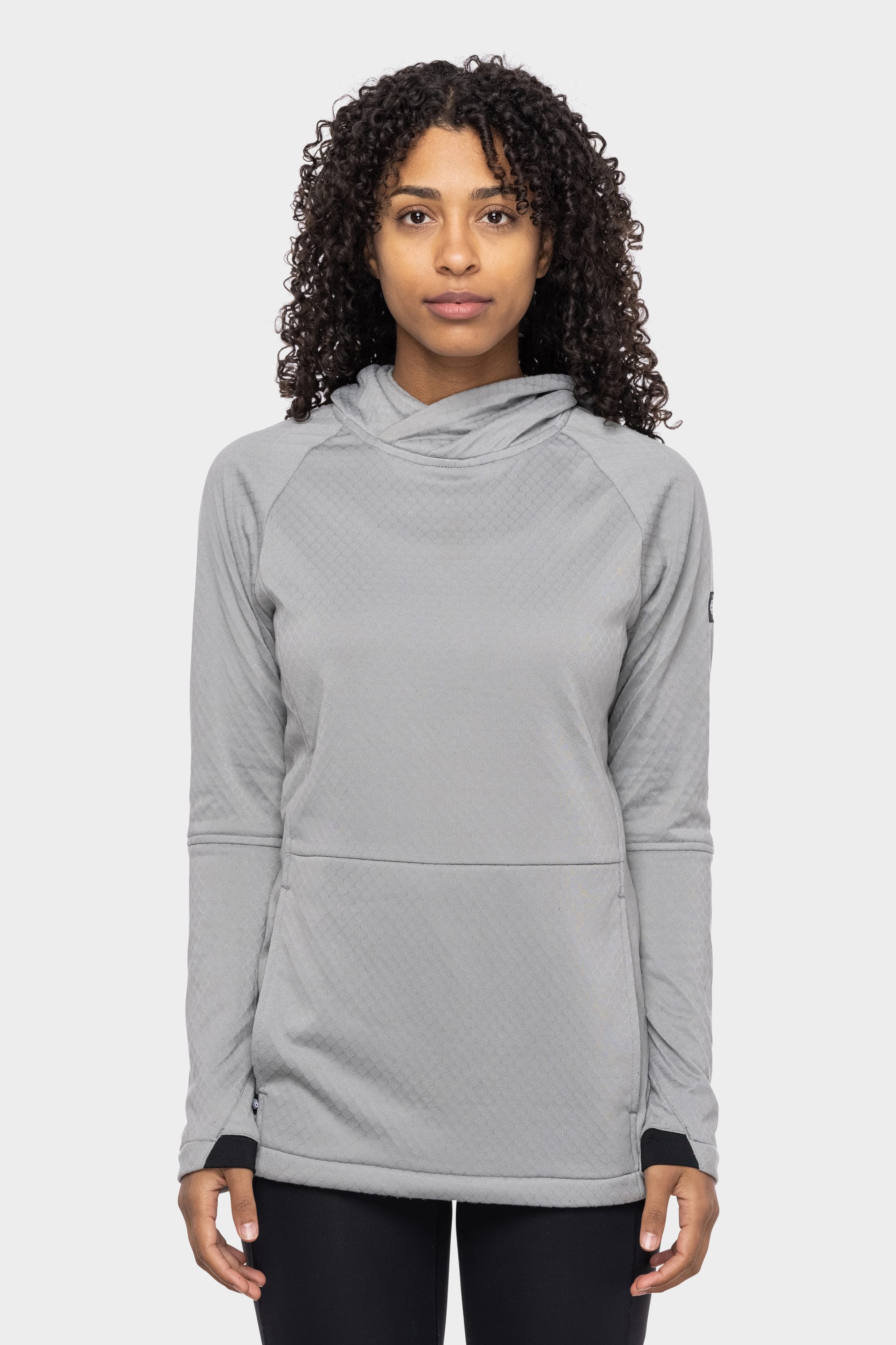 A person with curly hair wears the 686 Women's Mission Gridª Fleece Hoody in light gray, featuring a high collar, standing against a plain light background and facing forward with a neutral expression.