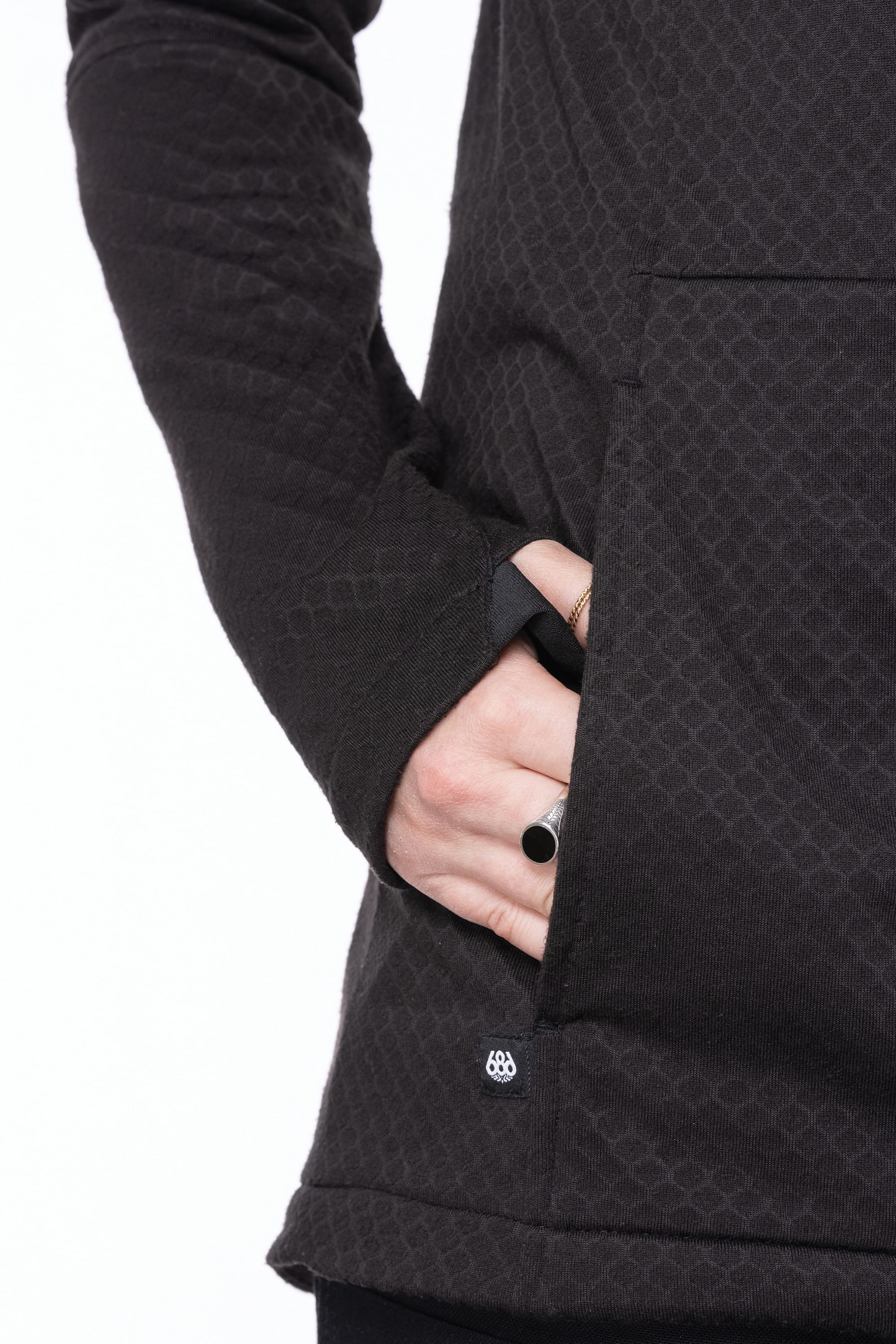 A person wears the 686 Women's Mission Gridª Fleece Hoody, featuring a textured black fabric and thumb-hole sleeves, with their right hand in the front pocket. They accessorize with two rings, including one with a black stone. Brand: 686.