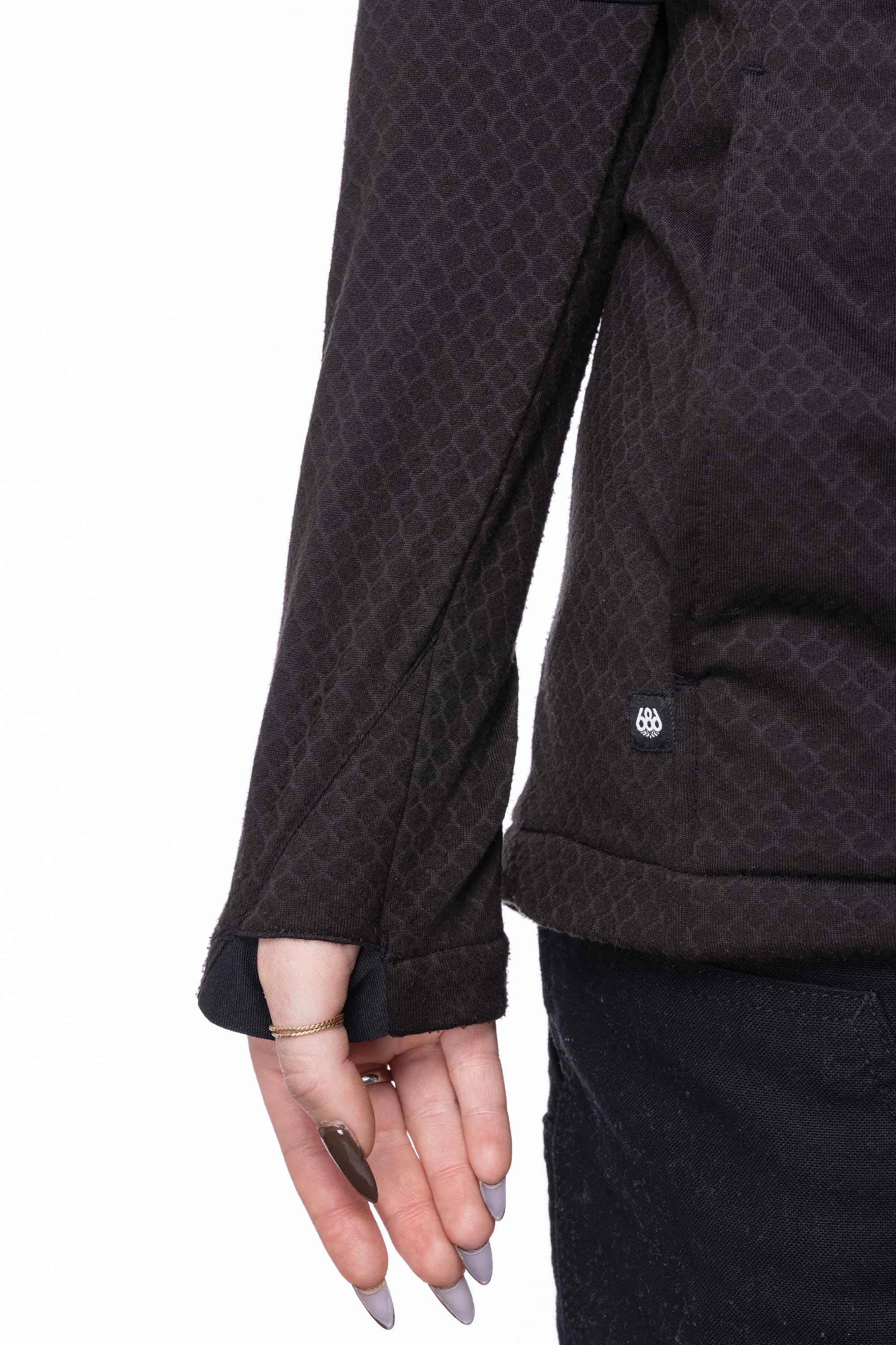 A woman models the 686 Women's Mission Gridª Fleece Hoody in black, featuring textured long sleeves with thumb holes, showcasing manicured nails and a delicate pinky ring against a white background.
