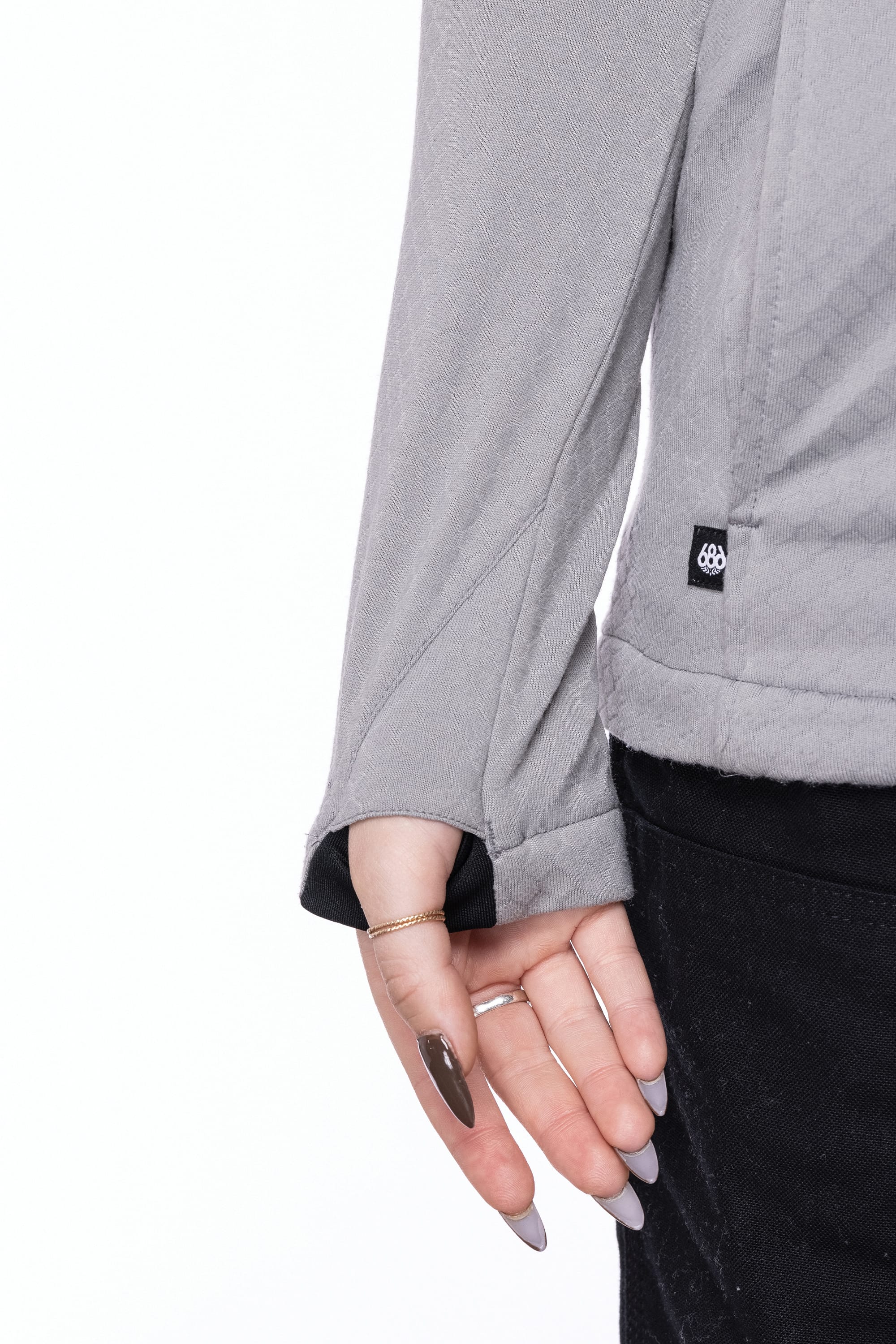 A person wears the 686 Women's Mission Gridª Fleece Hoody in gray, paired with black pants, revealing manicured nails and a thin gold ring as they gently pull at the sleeve near the hem.