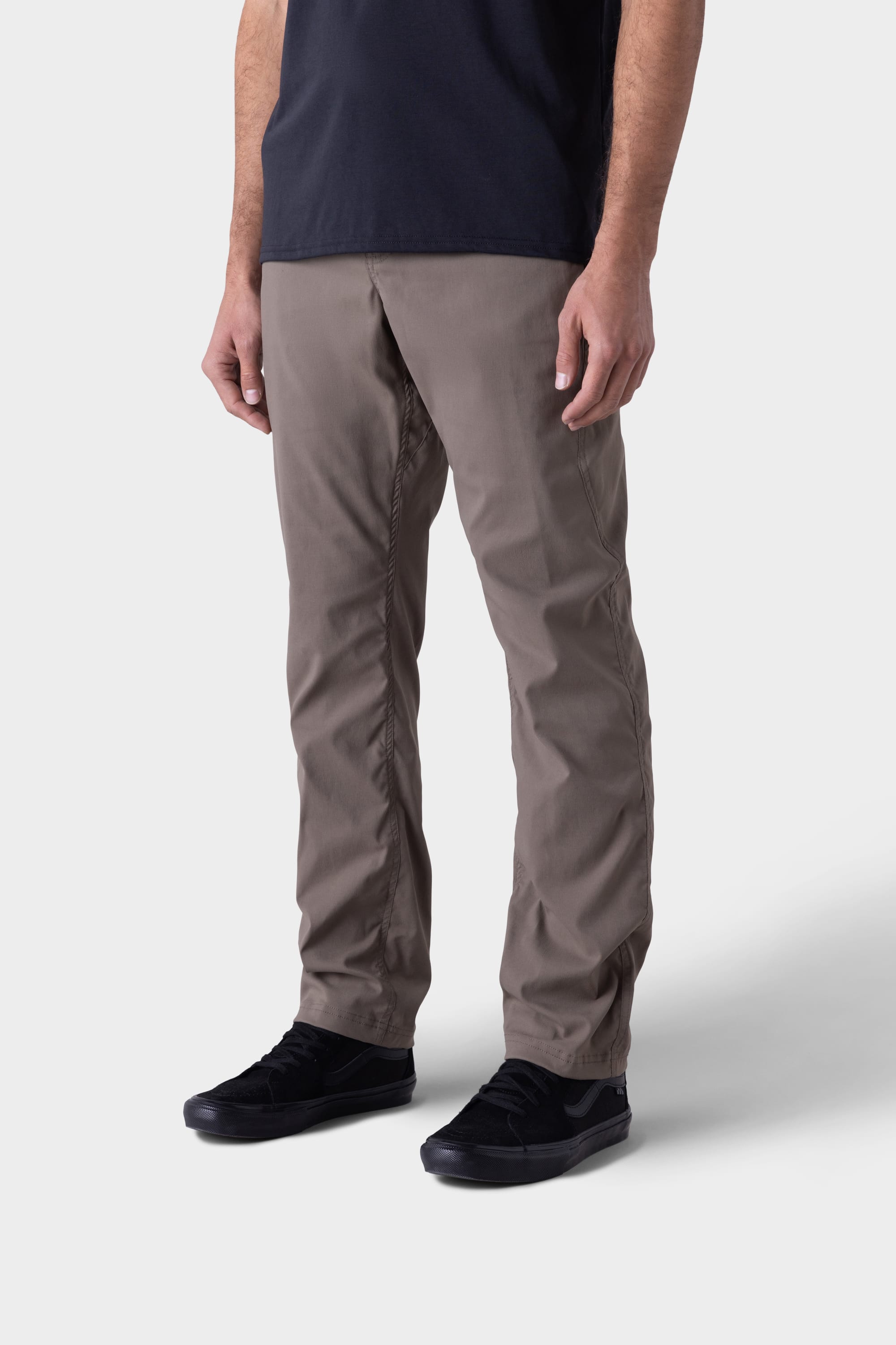 A man wearing the 686 Mens Platform Bike Pant - Relaxed Fit in taupe stands against a plain light gray background, shown from shoulders to feet, paired with a black T-shirt and black sneakers.