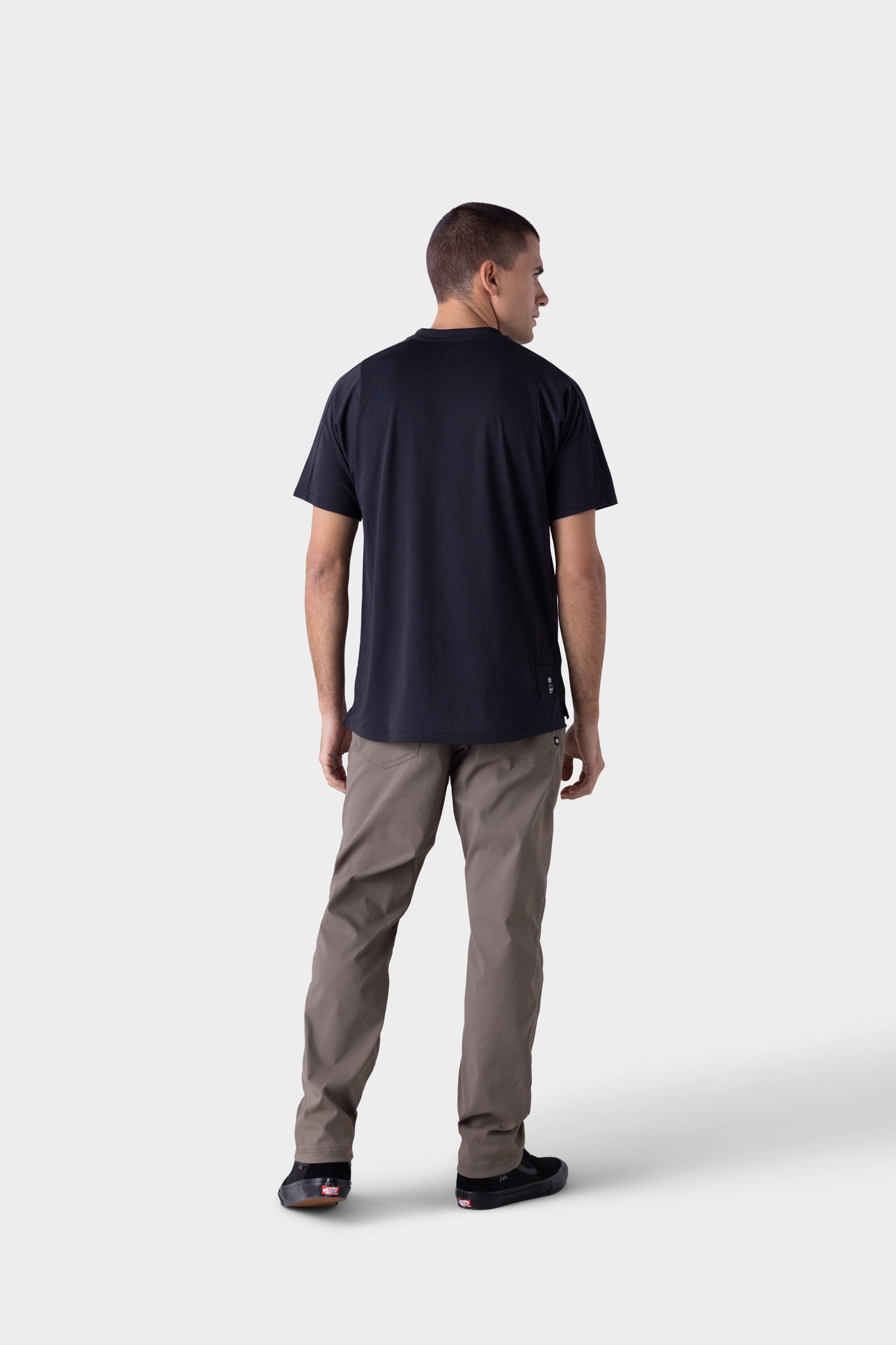 A man faces away from the camera, wearing 686 Mens Platform Bike Pant - Relaxed Fit in light brown, a black t-shirt, and black shoes against a plain light gray background.