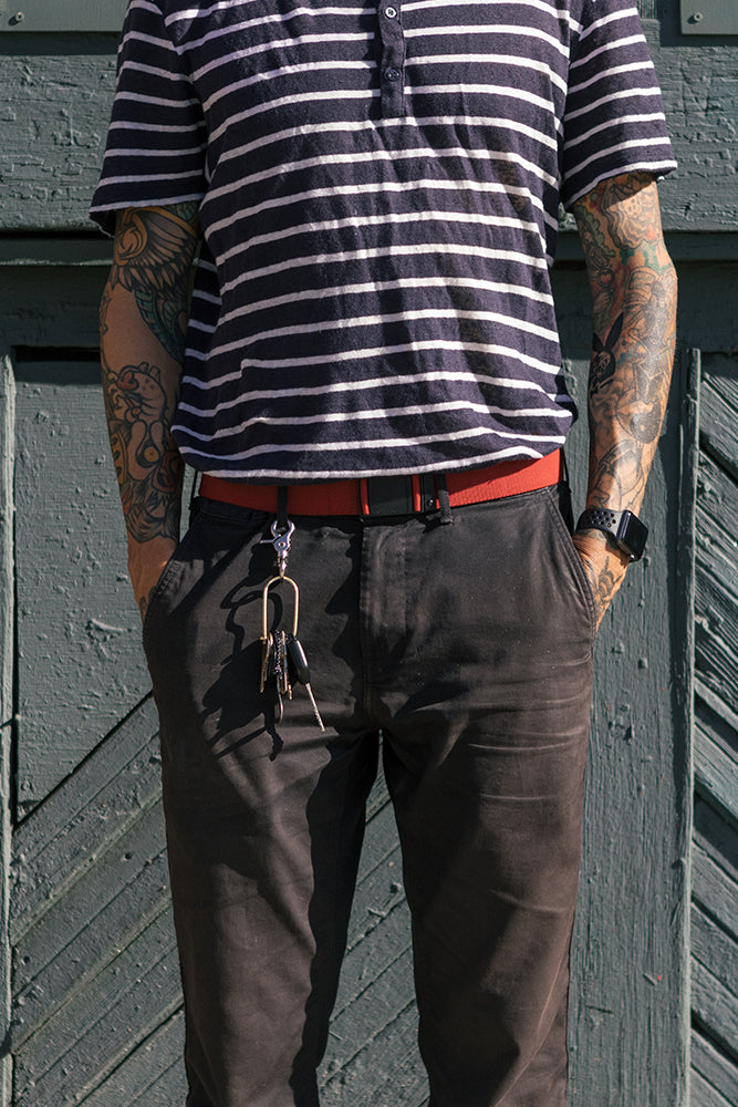 A person with tattooed arms stands against a green wooden wall, wearing a black and white striped shirt, black pants, the 686 Westwell Clicker Stretch Belt by 686 with keys clipped to it, and a black watch. Their hands are in their pockets.