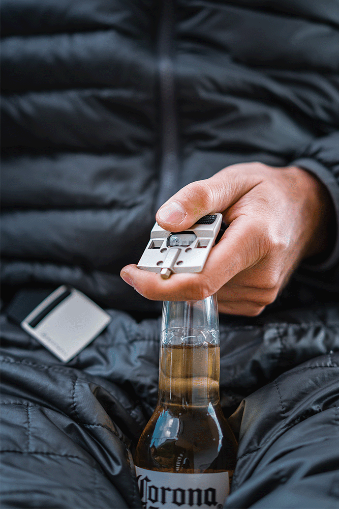 A person wearing a dark puffer jacket uses the 686 Westwell Clicker Stretch Belt by 686, which features a silver bottle opener on the buckle, to open a Corona beer bottle.