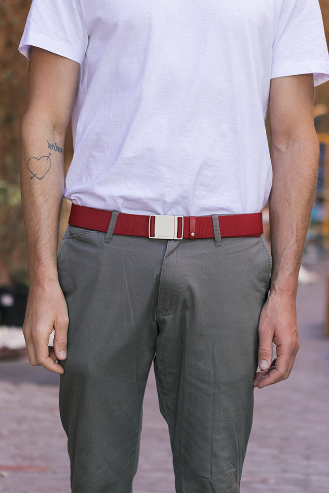 A person stands outdoors with arms at their sides, showing a simple heart tattoo on their left forearm. They wear a white t-shirt, gray pants, and the 686 Westwell Clicker Stretch Belt by 686. The photo is cropped at the shoulders.