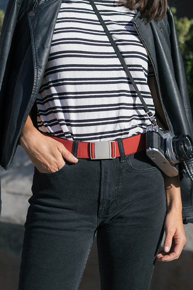 A person stands outdoors wearing a black leather jacket, black pants, a white-and-black striped shirt, and a red 686 Westwell Clicker Stretch Belt by 686, with a camera strapped across their shoulder.